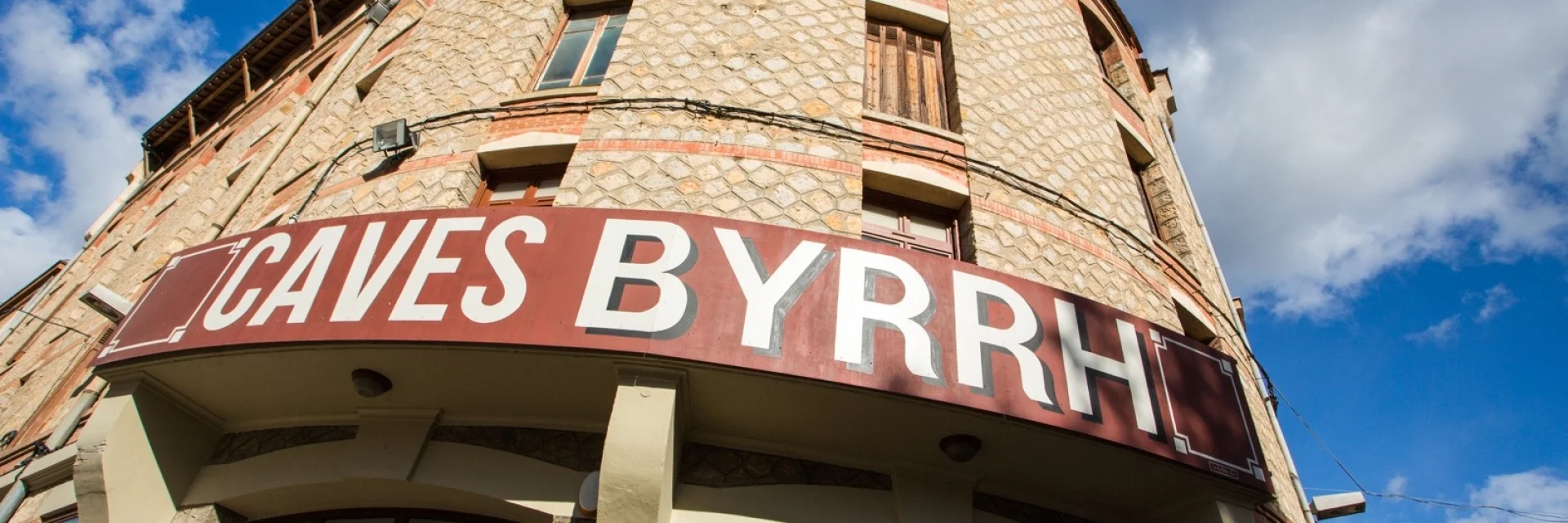 Façade en pierre des Caves Byrrh à Thuir, surmontée d’une grande enseigne rouge et blanche sous un ciel bleu parsemé de nuages.