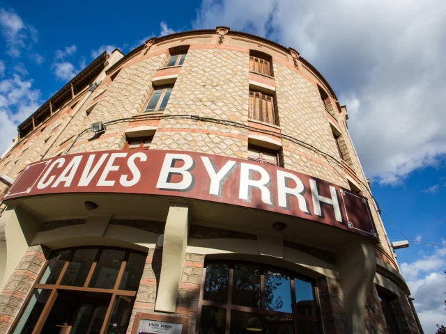 Façade en pierre des Caves Byrrh à Thuir, surmontée d’une grande enseigne rouge et blanche sous un ciel bleu parsemé de nuages.