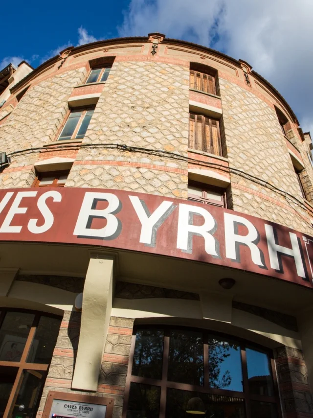 Façade en pierre des Caves Byrrh à Thuir, surmontée d’une grande enseigne rouge et blanche sous un ciel bleu parsemé de nuages.