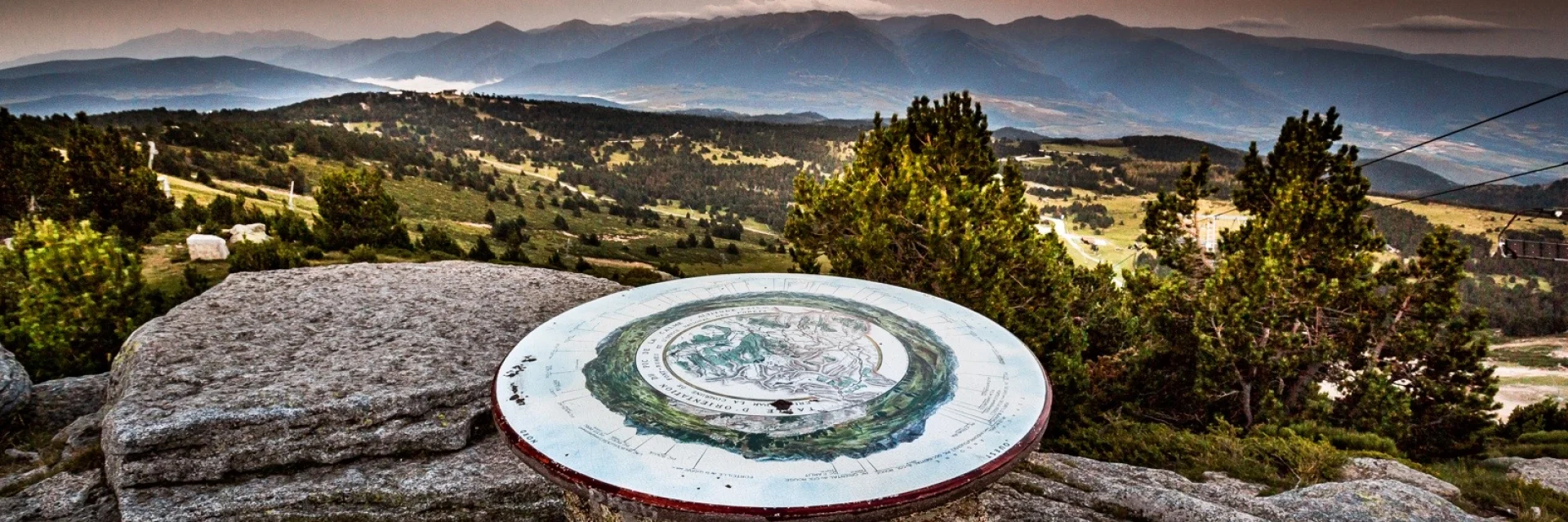 Table d’orientation en pierre sur un promontoire de montagne, offrant une vue panoramique sur les reliefs et vallées des Pyrénées-Orientales au coucher du soleil.