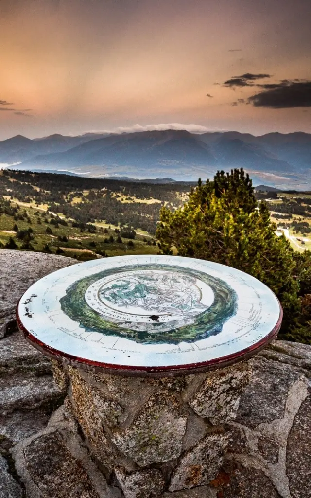 Table d’orientation en pierre sur un promontoire de montagne, offrant une vue panoramique sur les reliefs et vallées des Pyrénées-Orientales au coucher du soleil.