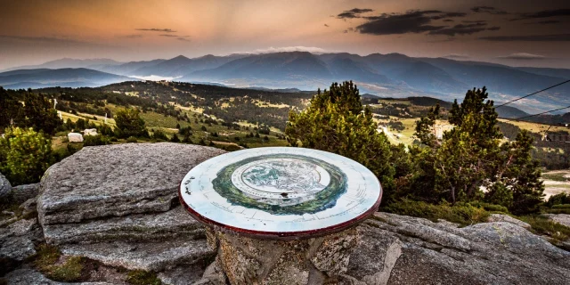 Table d’orientation en pierre sur un promontoire de montagne, offrant une vue panoramique sur les reliefs et vallées des Pyrénées-Orientales au coucher du soleil.