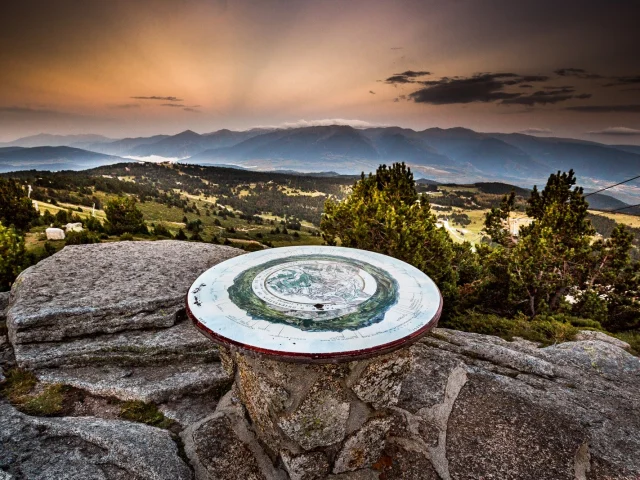 Table d’orientation en pierre sur un promontoire de montagne, offrant une vue panoramique sur les reliefs et vallées des Pyrénées-Orientales au coucher du soleil.
