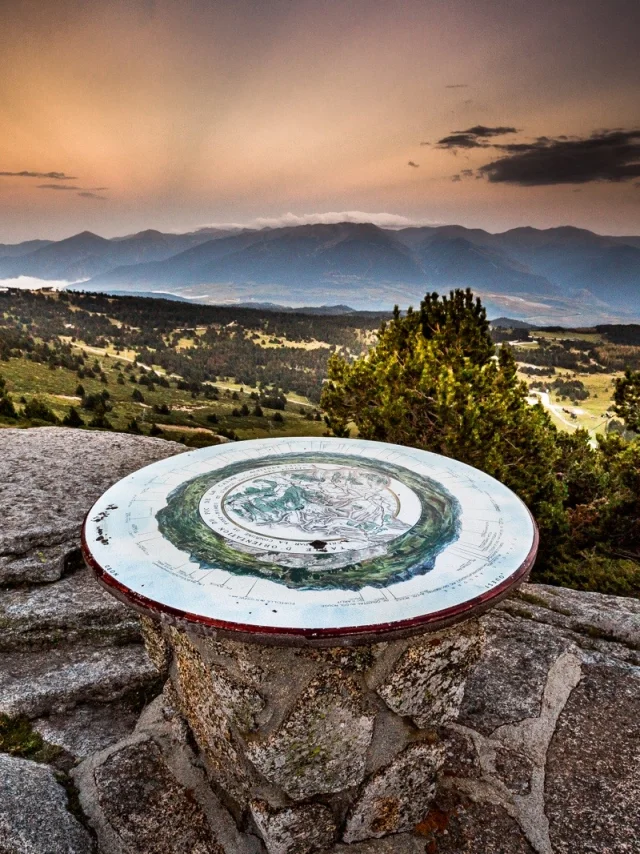 Table d’orientation en pierre sur un promontoire de montagne, offrant une vue panoramique sur les reliefs et vallées des Pyrénées-Orientales au coucher du soleil.