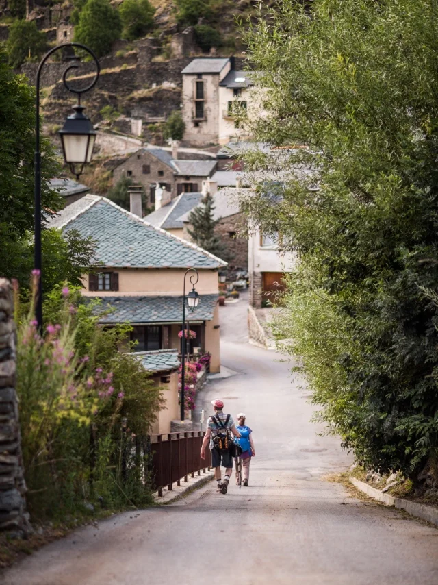 Deux randonneurs descendant une ruelle bordée de maisons en pierre dans un village de montagne des Pyrénées-Orientales.
