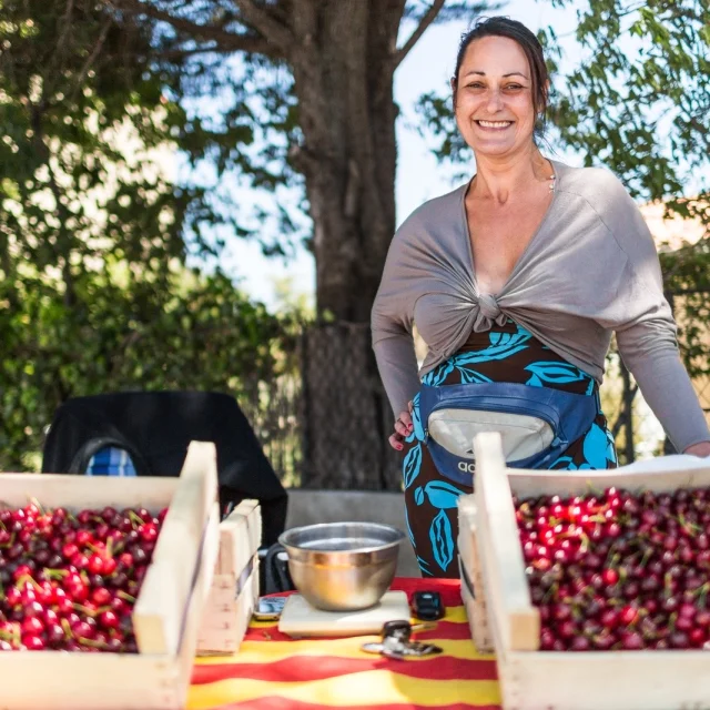Productrice souriante tenant un stand de cerises sur un marché en plein air, avec des cagettes remplies de fruits rouges fraîchement cueillis.