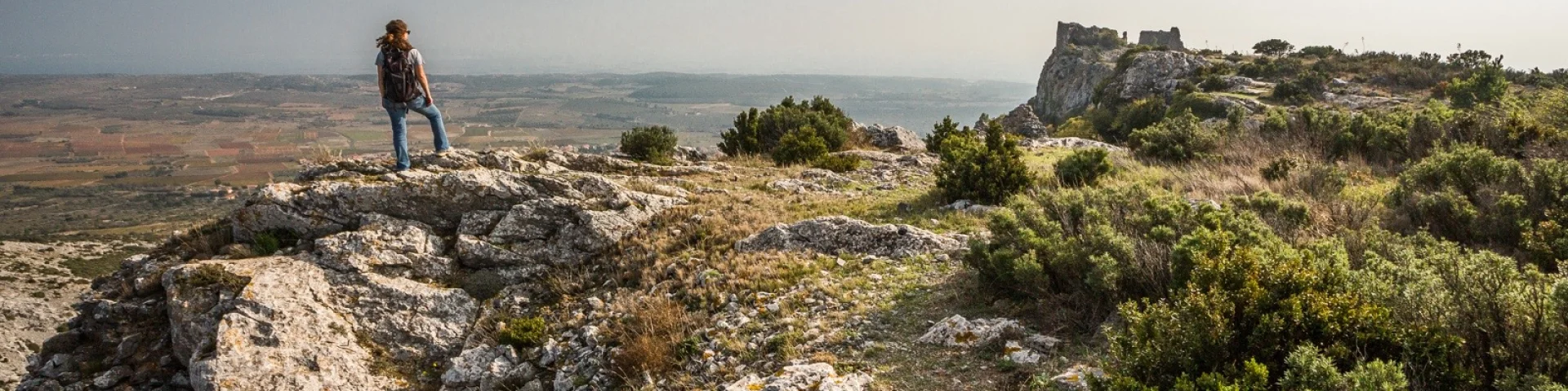 Randonneur sur un promontoire rocheux dominant la plaine du Roussillon, face aux ruines du château d’Opoul-Périllos sous un ciel légèrement voilé.