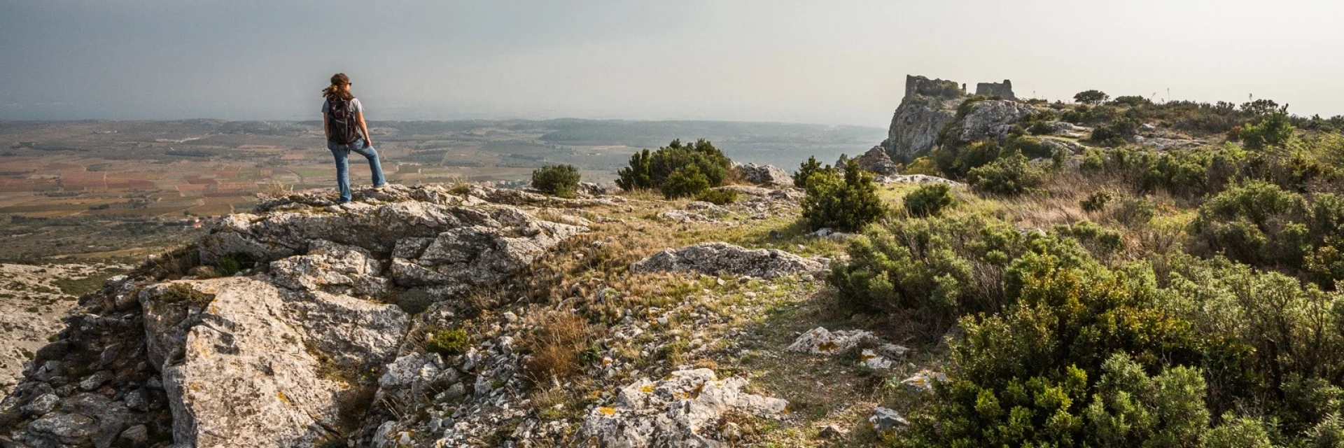 Randonneur sur un promontoire rocheux dominant la plaine du Roussillon, face aux ruines du château d’Opoul-Périllos sous un ciel légèrement voilé.