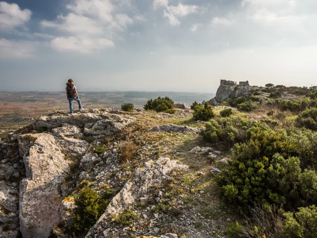 Randonneur sur un promontoire rocheux dominant la plaine du Roussillon, face aux ruines du château d’Opoul-Périllos sous un ciel légèrement voilé.