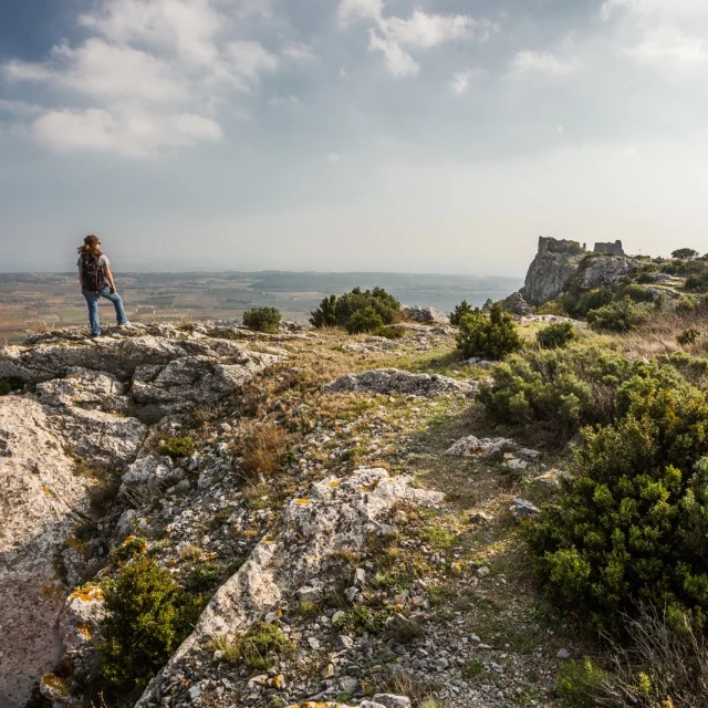 Randonneur sur un promontoire rocheux dominant la plaine du Roussillon, face aux ruines du château d’Opoul-Périllos sous un ciel légèrement voilé.