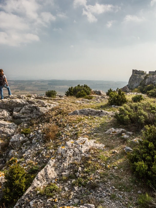 Randonneur sur un promontoire rocheux dominant la plaine du Roussillon, face aux ruines du château d’Opoul-Périllos sous un ciel légèrement voilé.