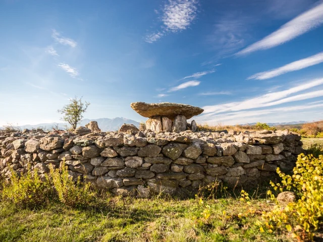 Dolmen en pierre sèche entouré d’un muret circulaire, sous un ciel bleu strié de nuages, avec les montagnes en arrière-plan.