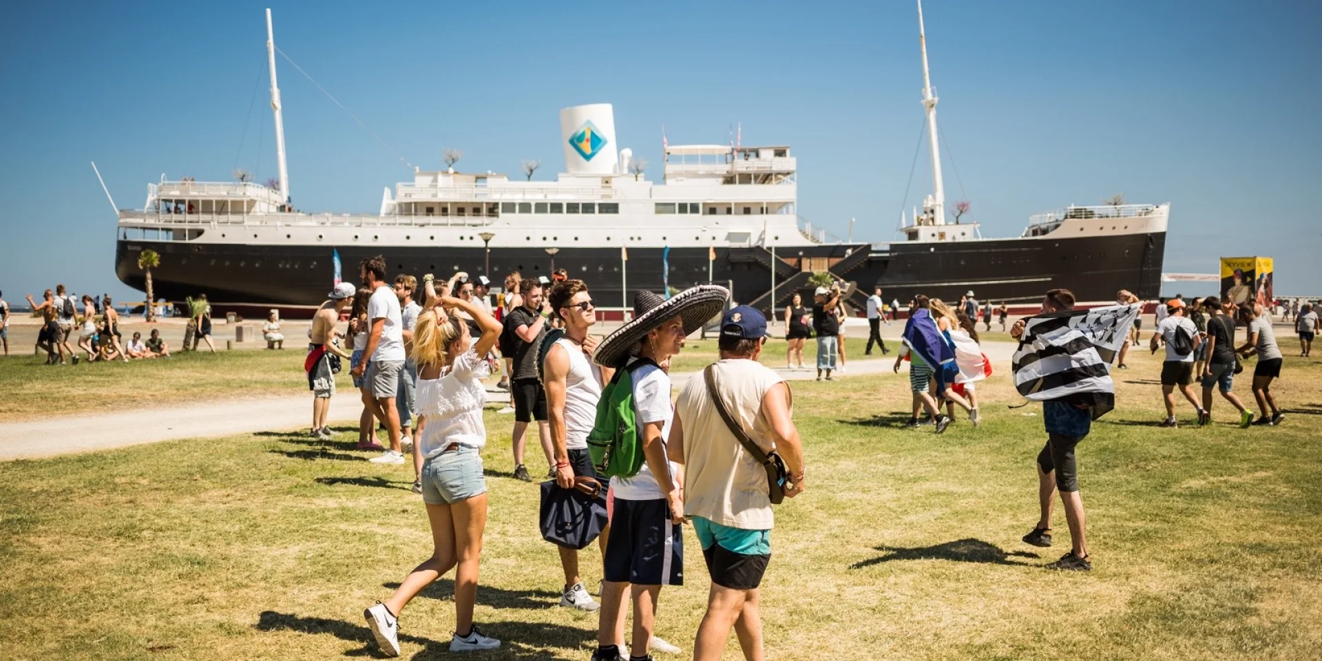 Groupe de visiteurs marchant sur la pelouse face au paquebot Lydia, emblème du Barcarès, sous un ciel bleu d’été.