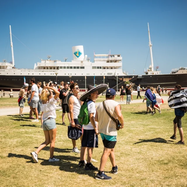 Groupe de visiteurs marchant sur la pelouse face au paquebot Lydia, emblème du Barcarès, sous un ciel bleu d’été.