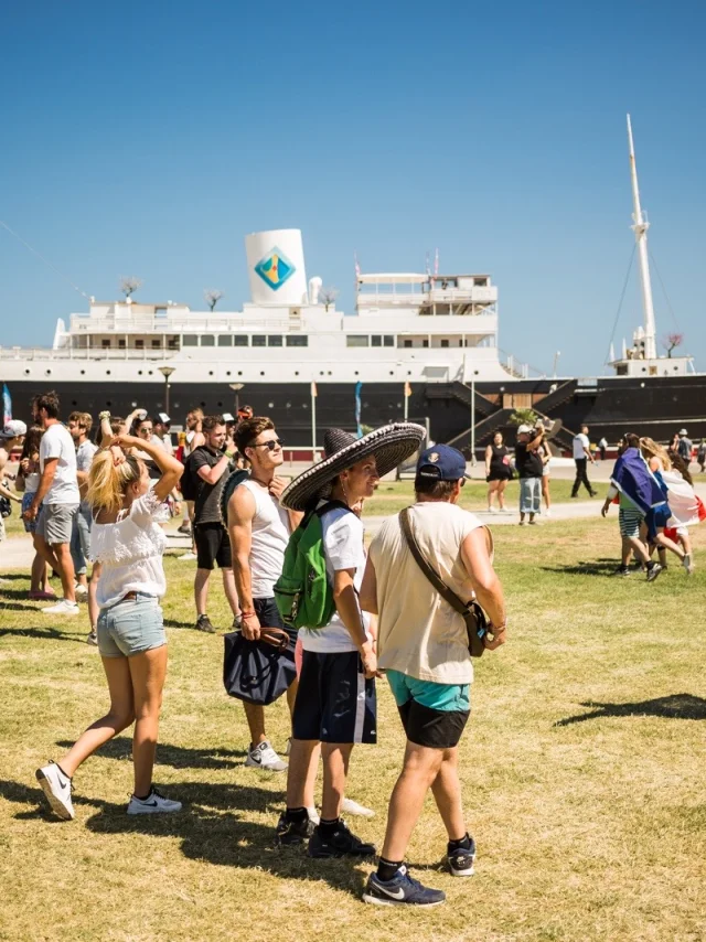 Groupe de visiteurs marchant sur la pelouse face au paquebot Lydia, emblème du Barcarès, sous un ciel bleu d’été.