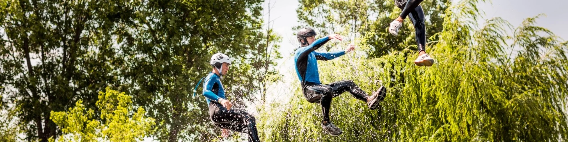 Trois personnes en combinaison et casque sautant en même temps d’une plateforme au-dessus de l’eau lors d’une activité aquatique de plein air.