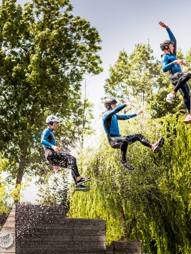 Trois personnes en combinaison et casque sautant en même temps d’une plateforme au-dessus de l’eau lors d’une activité aquatique de plein air.