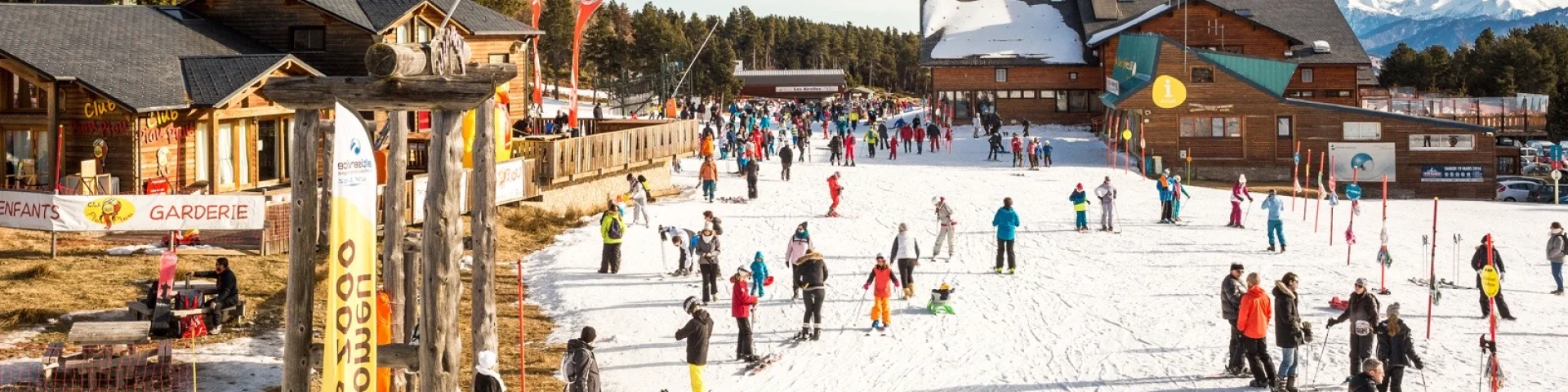 FNombreux skieurs et familles sur le front de neige de Font-Romeu, entouré de chalets en bois et de montagnes enneigées sous un ciel bleu.