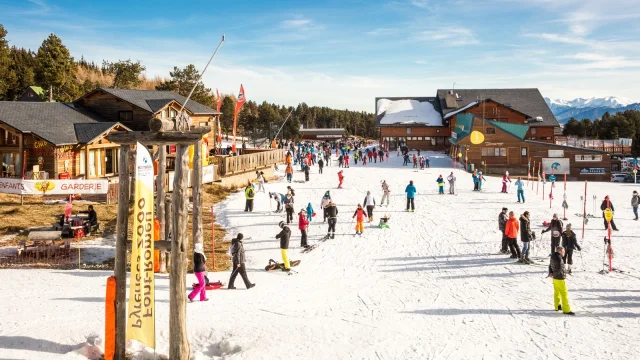 FNombreux skieurs et familles sur le front de neige de Font-Romeu, entouré de chalets en bois et de montagnes enneigées sous un ciel bleu.