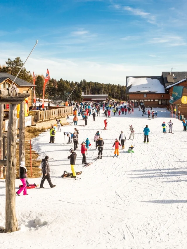 FNombreux skieurs et familles sur le front de neige de Font-Romeu, entouré de chalets en bois et de montagnes enneigées sous un ciel bleu.