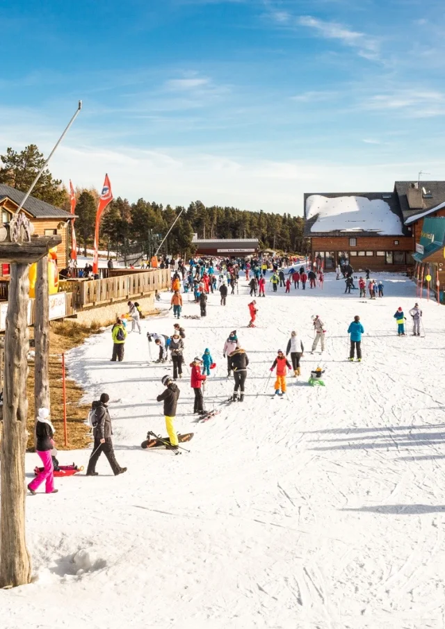 FNombreux skieurs et familles sur le front de neige de Font-Romeu, entouré de chalets en bois et de montagnes enneigées sous un ciel bleu.