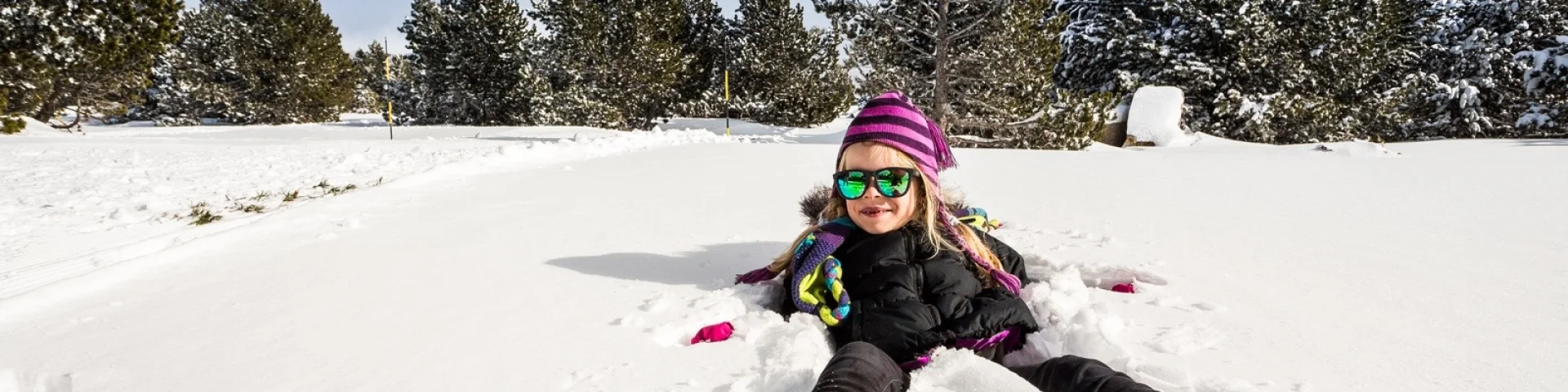 Enfant souriant allongé dans la neige, portant bonnet violet et lunettes de soleil, entouré d’arbres enneigés sous un ciel clair.