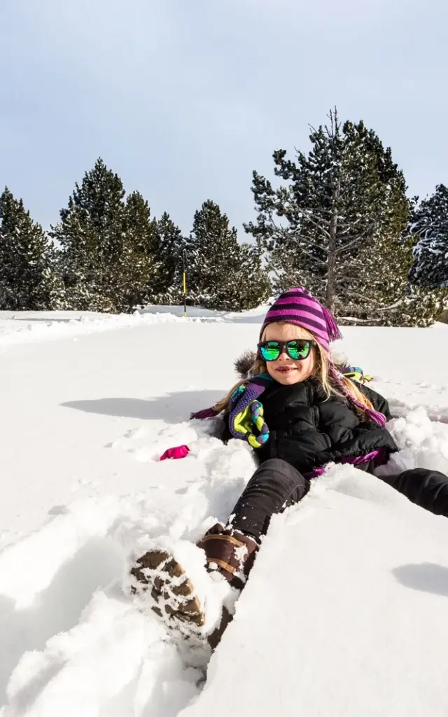 Enfant souriant allongé dans la neige, portant bonnet violet et lunettes de soleil, entouré d’arbres enneigés sous un ciel clair.