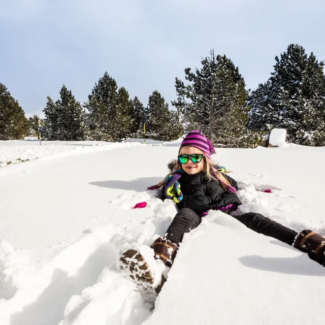Enfant souriant allongé dans la neige, portant bonnet violet et lunettes de soleil, entouré d’arbres enneigés sous un ciel clair.
