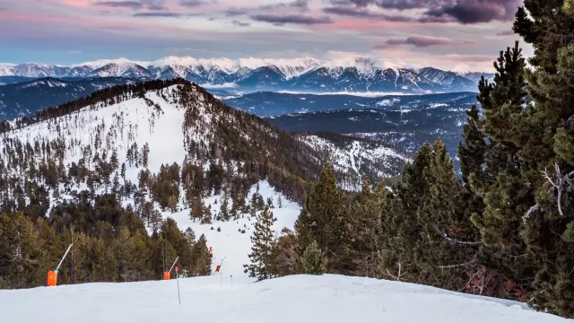 Panorama hivernal sur les montagnes enneigées des Pyrénées-Orientales, avec une piste de ski bordée de pins et un ciel coloré au coucher du soleil.