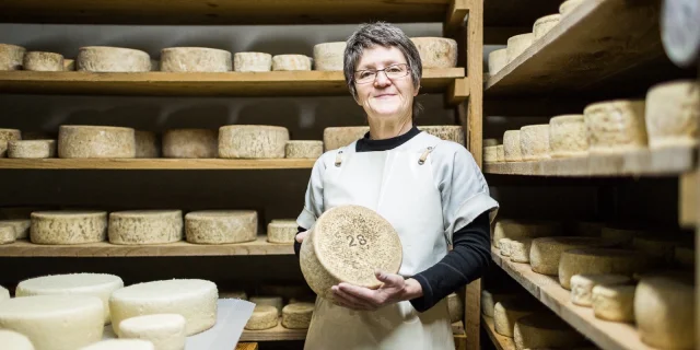 Fromagère souriante tenant une meule de fromage dans une cave d’affinage aux étagères en bois remplies de fromages.