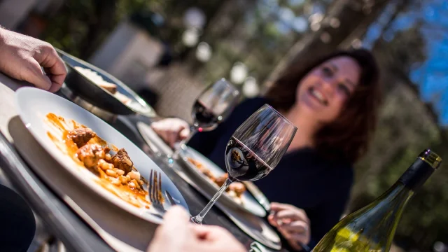 Repas convivial en terrasse avec deux personnes partageant un plat catalan et un verre de vin rouge sous le soleil.
