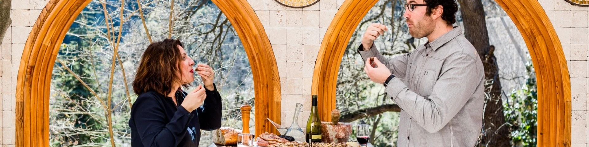 Deux personnes dégustent des produits du terroir catalan autour d’une table en pierre, avec vin, charcuterie et pain, dans un cadre lumineux aux arches en bois ouvrant sur la nature.