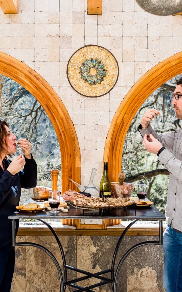Deux personnes dégustent des produits du terroir catalan autour d’une table en pierre, avec vin, charcuterie et pain, dans un cadre lumineux aux arches en bois ouvrant sur la nature.