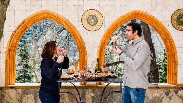 Deux personnes dégustent des produits du terroir catalan autour d’une table en pierre, avec vin, charcuterie et pain, dans un cadre lumineux aux arches en bois ouvrant sur la nature.