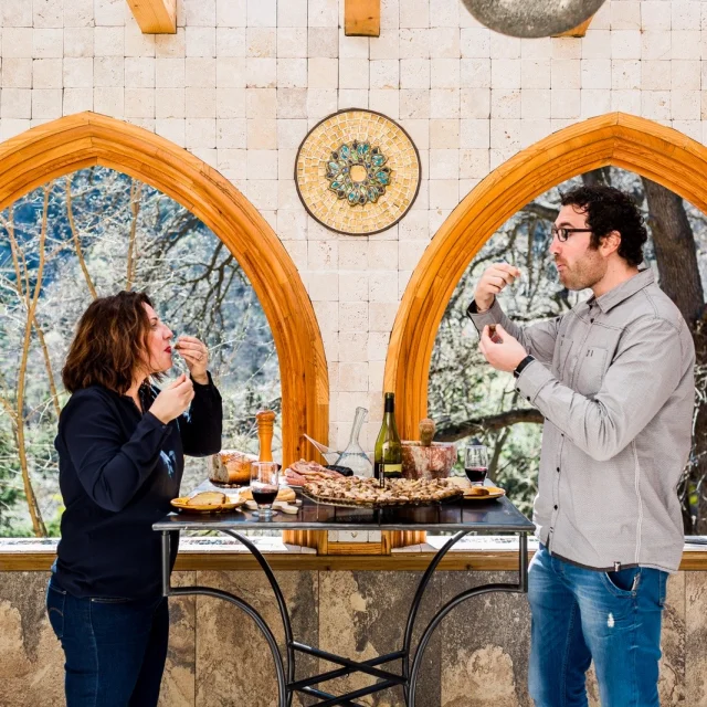Deux personnes dégustent des produits du terroir catalan autour d’une table en pierre, avec vin, charcuterie et pain, dans un cadre lumineux aux arches en bois ouvrant sur la nature.