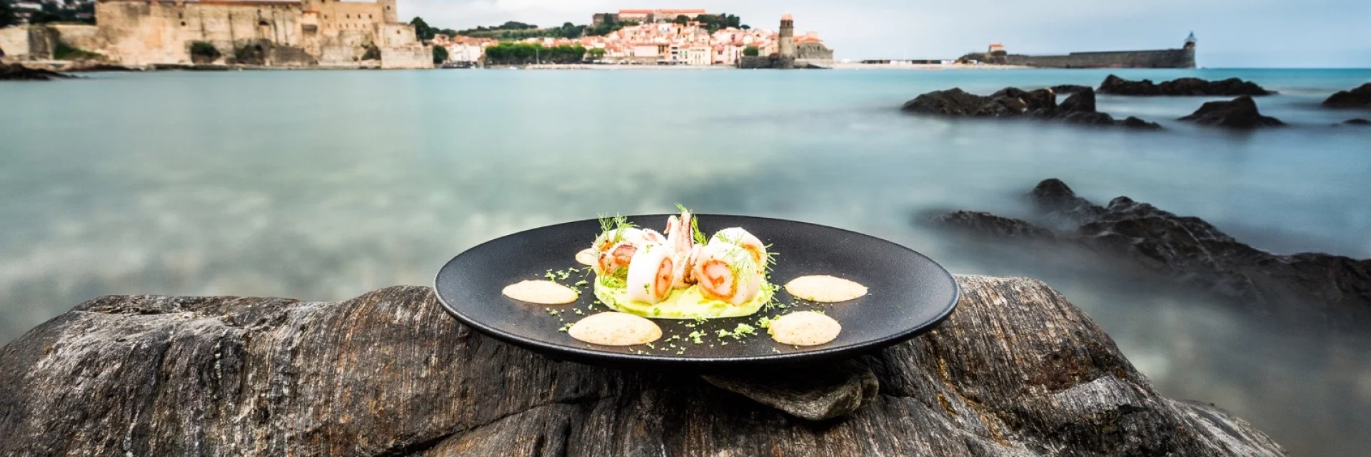Assiette gastronomique dressée sur un rocher face à la baie de Collioure, avec vue sur le Château Royal et l’église Notre-Dame-des-Anges.