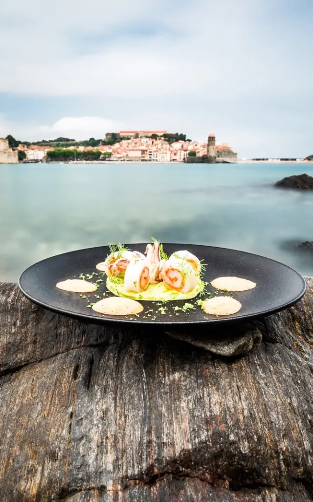 Assiette gastronomique dressée sur un rocher face à la baie de Collioure, avec vue sur le Château Royal et l’église Notre-Dame-des-Anges.