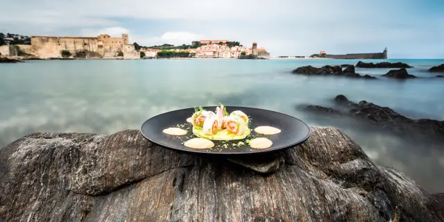 Assiette gastronomique dressée sur un rocher face à la baie de Collioure, avec vue sur le Château Royal et l’église Notre-Dame-des-Anges.