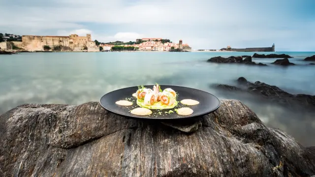 Assiette gastronomique dressée sur un rocher face à la baie de Collioure, avec vue sur le Château Royal et l’église Notre-Dame-des-Anges.