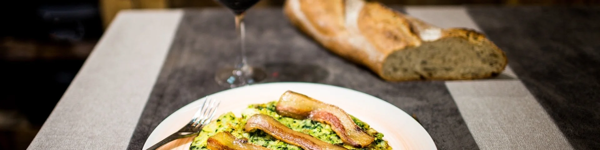 Assiette de trinxat catalan, galette de pommes de terre et de chou surmontée de tranches de lard grillé, servie avec un verre de vin rouge et du pain sur la table.