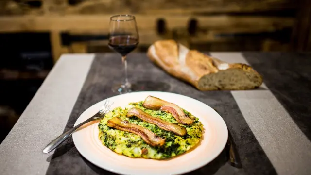 Assiette de trinxat catalan, galette de pommes de terre et de chou surmontée de tranches de lard grillé, servie avec un verre de vin rouge et du pain sur la table.