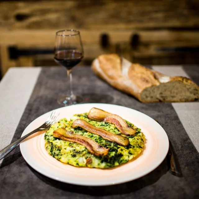Assiette de trinxat catalan, galette de pommes de terre et de chou surmontée de tranches de lard grillé, servie avec un verre de vin rouge et du pain sur la table.