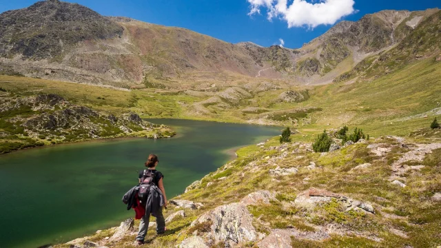 Randonneuse en montagne longeant un lac d’altitude aux eaux vert émeraude, entouré de sommets rocheux sous un ciel bleu.