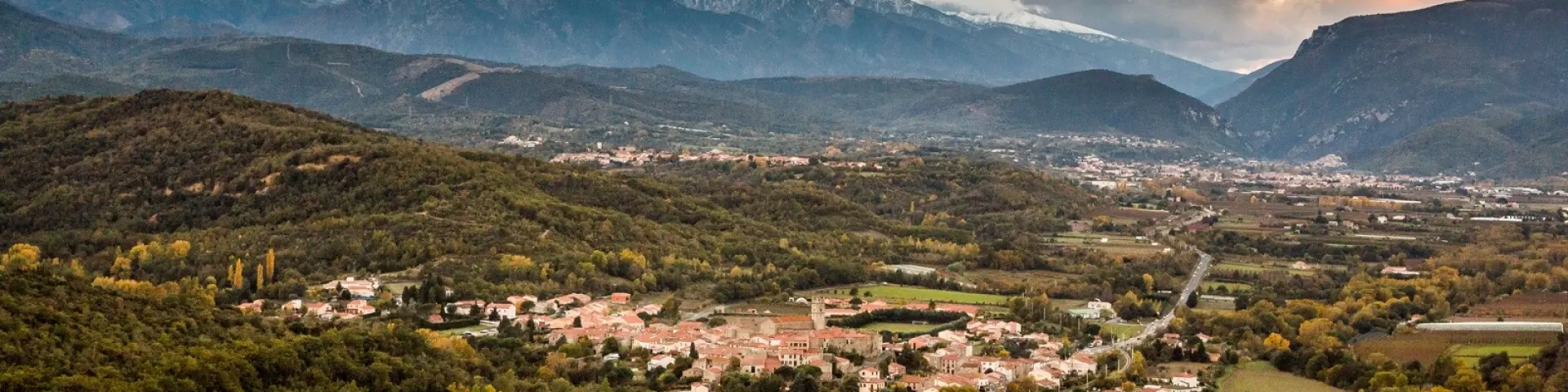 Vue sur Marquixanes depuis Arboussols. Vue panoramique d’un village niché dans une vallée verdoyante au pied des montagnes, sous un ciel nuageux au coucher du soleil.