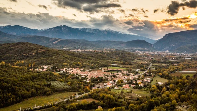 Vue sur Marquixanes depuis Arboussols. Vue panoramique d’un village niché dans une vallée verdoyante au pied des montagnes, sous un ciel nuageux au coucher du soleil.