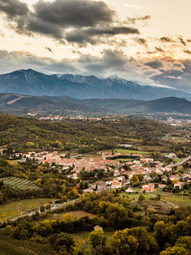 Vue sur Marquixanes depuis Arboussols. Vue panoramique d’un village niché dans une vallée verdoyante au pied des montagnes, sous un ciel nuageux au coucher du soleil.