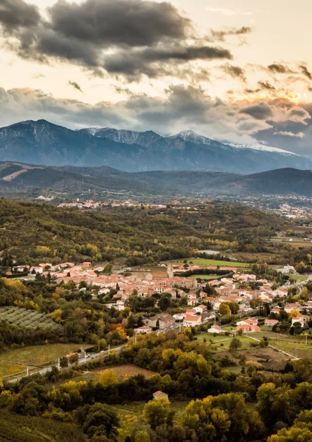 Vue sur Marquixanes depuis Arboussols. Vue panoramique d’un village niché dans une vallée verdoyante au pied des montagnes, sous un ciel nuageux au coucher du soleil.