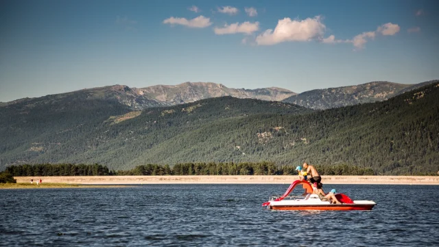 Famille en pédalo rouge avec toboggan sur le lac de Matemale, entouré des montagnes boisées du Capcir sous un ciel bleu d’été.