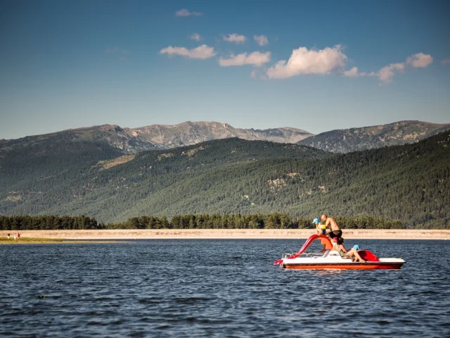Famille en pédalo rouge avec toboggan sur le lac de Matemale, entouré des montagnes boisées du Capcir sous un ciel bleu d’été.