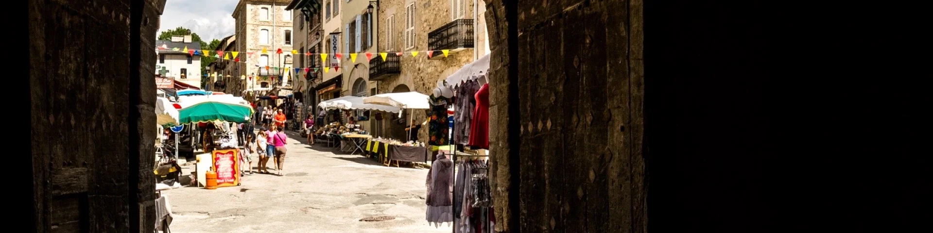 Passage voûté menant à la rue commerçante de Mont-Louis animée, avec ses étals, ses promeneurs et ses façades en pierre typiques d’une cité fortifiée.